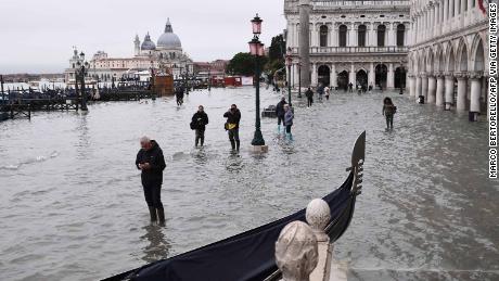 People walk past a stranded gondola across the flooded Riva degli Schiavoni embankment and St. Mark's Square in the background.