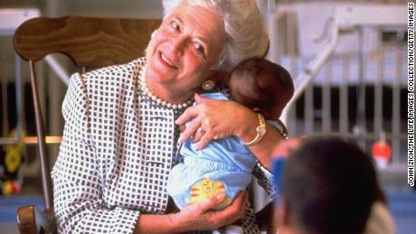 First Lady Barbara Bush holding baby while two-year-old child takes a photo wtih a toy camera at hospice for children with AIDS. 