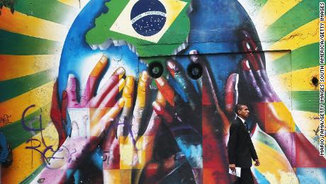 ชายคนหนึ่งผ่านกราฟฟิตีของมือหลากสีที่รองรับดาวเคราะห์ที่มีธงชาติบราซิลเมื่อวันที่ 10 มิถุนายน 2014 ในเมืองเซาเปาโล ประเทศบราซิล  แมตช์แรกสำหรับฟุตบอลโลก 2014 คือวันที่ 12 มิถุนายน ที่เซาเปาโล เมื่อบราซิลพบกับโครเอเชีย
