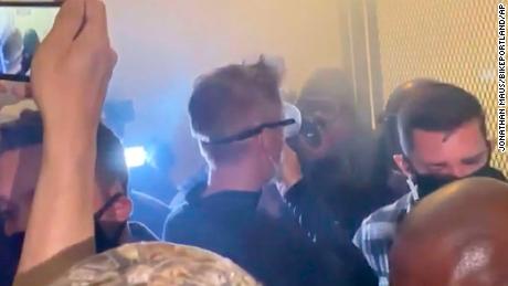 In this image made from video released by Jonathan Maus, Portland Mayor Ted Wheeler, center in black with goggles looking away, stands at a fence guarding a federal courthouse as tear gas drifts by early July 23, 2020, in Portland Oregon.