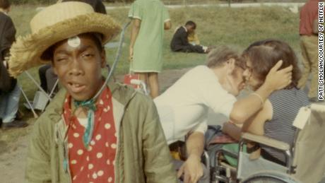 Caption:, Camper Cary Walker poses with a straw hat on, while in the background, a couple in wheelchairs shares a romantic embrace., Visual Description:, Vintage color snapshot at Camp Jened. Cary Walker, a young African American camper, squints at the camera with a bemused look, wearing a straw hat, a red shirt with white-polka dots, and an army-green jacket. He appears to have a white sticker on his forehead. Behind him, a couple sitting in wheelchairs share a romantic embrace, looking like they just kissed or are about to kiss., Alt Text:, Vintage snapshot of two campers in wheelchairs kissing while another camper squints at the camera wearing a straw hat.