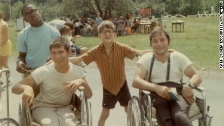 Caption:, 1968 outdoor color snapshot. Four male campers, two in wheelchairs, smile for the camera. Behind them a large group assembles in the Jened field., Visual Description:, two sitting in manual wheelchairs and two standing, smile for the camera. The boy standing in the middle has his arms on the backs of the wheelchairs to either side of him. Behind them a large group assembles in the Camp Jened field., Alt Text:, Vintage snapshot of four Jened campers, smiling for the camera.