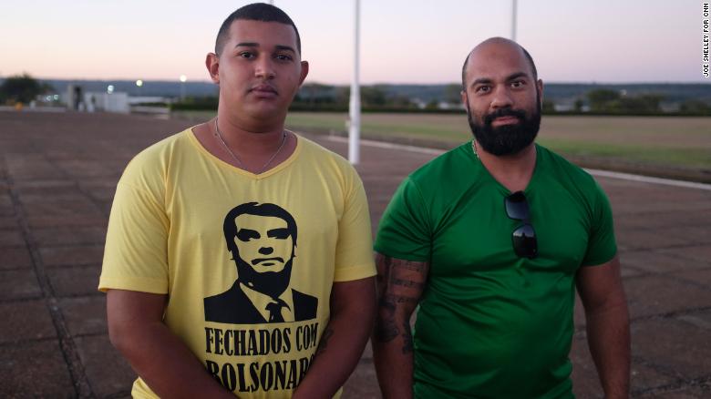 Supporters of President Bolsonaro gather outside his residence. One wears a t-shirt that reads &quot;Sticking with Bolsonaro&quot; on the front and &quot;All power to the people&quot; on the back.