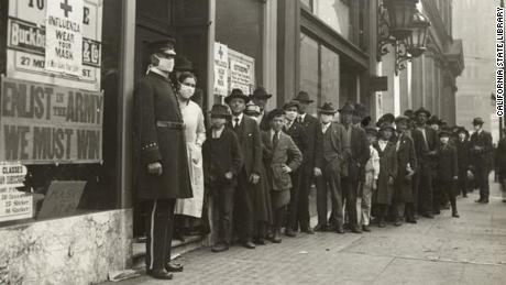 A line of people waiting for flu masksin 1918 in San Francisco. A sign on the window bearing the Red Cross logo reads: &quot;Influenza. Wear Your Mask.&quot;