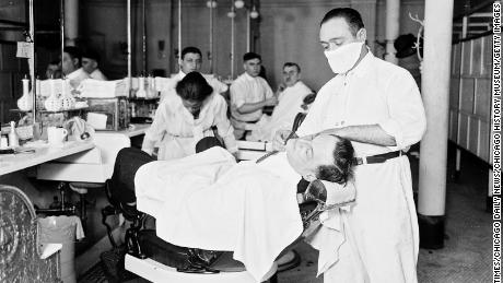 Un frizer care purta o mască antigripală bărbiereste bărbatul în timpul unei pandemii în curs de desfășurare din Chicago, în 1918.