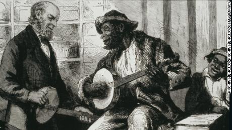 A Black man plays a banjo for White listeners in the 1880s United States. 