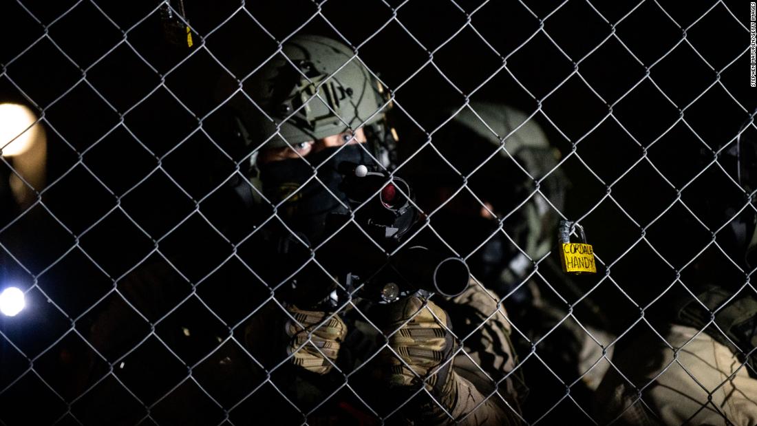 A law enforcement officer points a projectile weapon through fencing outside the police headquarters.