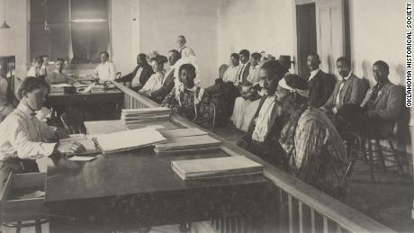Photograph of Chickasaw Freedmen filing on allotments at Tishomingo, Indian Territory. The freedmen are seated in two rows in front of large desks occupied by what appear to be clerks.