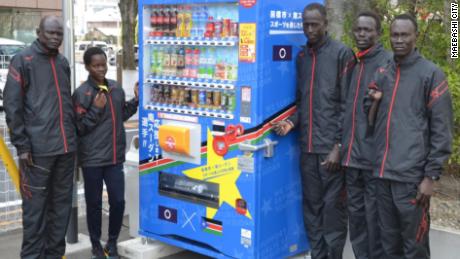 南スーダンのチームとコーチのジョセフ（左端）がメイバシの自動販売機の前で、自国の旗を掲げています。 
