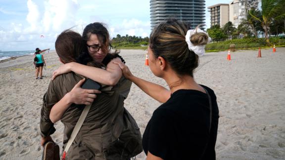 Faydah Bushnaq, center, is hugged by Maria Fernanda Martinez as they stand on the beach near the building. Bushnaq, who was vacationing in South Florida, stopped to write "pray for their souls" in the sand.