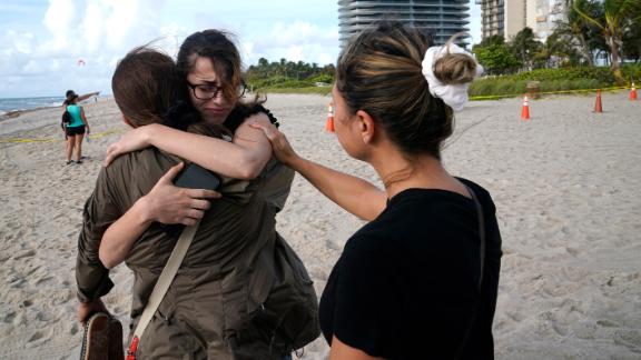 Faydah Bushnaq, center, is hugged by Maria Fernanda Martinez as they stand on the beach near the building. Bushnaq, who was vacationing in South Florida, stopped to write "pray for their souls" in the sand.