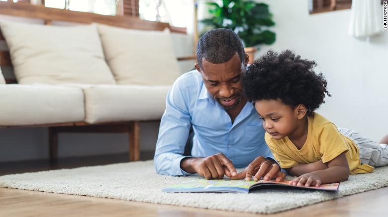 A father and his son read a book while lying on the floor at home.