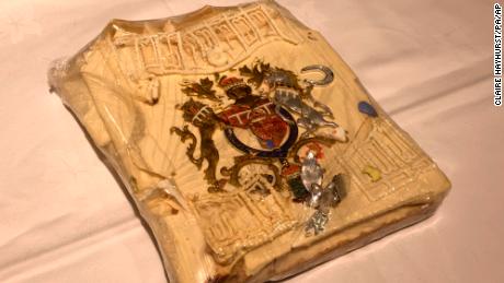 The cake slice features a colorful coat of arms in gold, red, blue and silver, a silver horseshoe and leaf spray, as well as white decorative frosting.