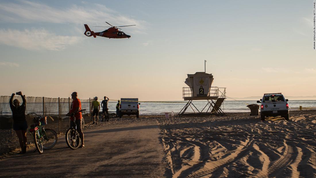A helicopter flies over bystanders in Huntington Beach. The federal Bureau of Safety and Environmental Enforcement is assisting in the Coast Guard-led response to the oil spill, the agency told CNN.