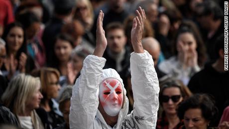 Протестующий в маске с изображением инъекций и полном защитном костюме аплодирует в Женеве 9 октября 2021 года.