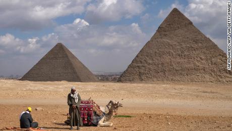 A picture taken on December 17, 2021 shows men resting next to a camel with the Great Pyramid of Khufu (Cheops) (L) and Pyramid of Khafre (Chephren) (R) in the background at the Giza pyramids necropolis on the southwestern outskirts of the Egyptian capital Cairo. (Photo by SAFIN HAMED / AFP) (Photo by SAFIN HAMED/AFP via Getty Images)