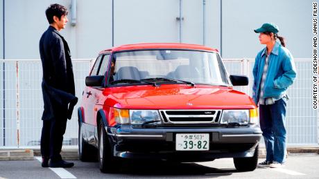Hidetoshi Nishijima and Toko Miura as Yusuke Kafuku and Misaki Watari standing next to the red Saab 900 Turbo in Hamaguchi&#39;s &quot;Drive My Car.&quot; 
