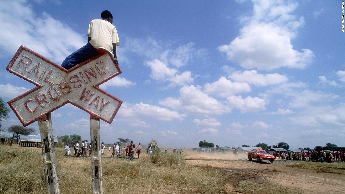 A spectator perched on a railway crossing sign watches Finnish driver Tommi Makinen in action in his Mitsubishi Lancer Evo VII during the 2001 Safari World Rally Championship.