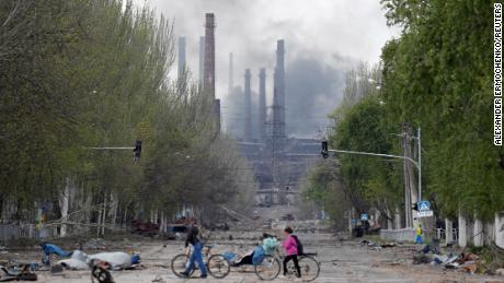La gente va in bicicletta per strada mentre il fumo sale sopra l'acciaieria Azovstal a Mariupol, in Ucraina, il 2 maggio. 