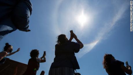 People watch a rainbow form a circle around the sun as they mourn and pray in the street outside Tops.