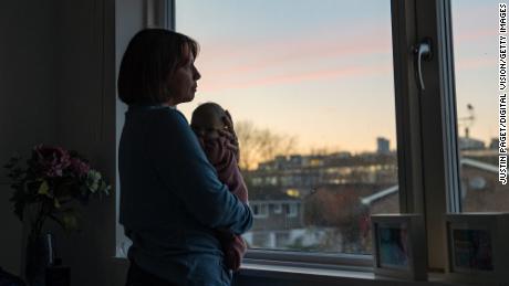 woman holds her baby to her chest as she looks through her bedroom window at sunset 