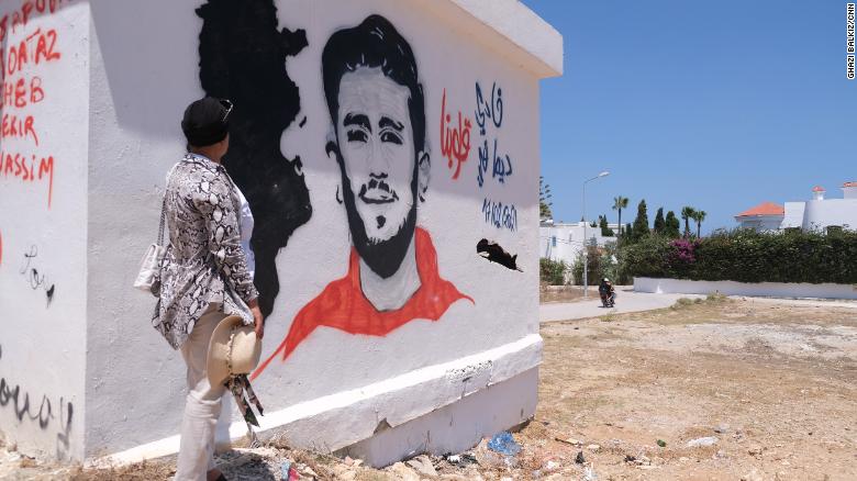 Samia se tient près d'une peinture murale de son fils dans leur ville natale de Kelibia.  Elle a dit que ses amis, qui lui rendent régulièrement visite, l'ont peinte.  Le dessin a son visage, la date de sa disparition, une carte de la Tunisie et dit "Fadi toujours dans nos cœurs".