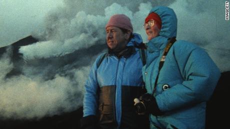 Katia and Maurice Krafft, in blue winter jackets, gaze upon a volcano in the distance as smoke, steam and ash swirl behind them. (Credit: Image&#39;Est)