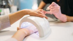 Female client dries gel varnish in the nail dryer, beautician in pink gloves on background, beauty salon. Manicurist doing hand care cosmetic procedure