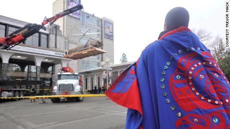An Indigenous tribal member wearing their clan&#39;s regalia, embroidered with their family crest, watches as the totem pole is lifted out of the Royal BC Museum and lowered to the ground.