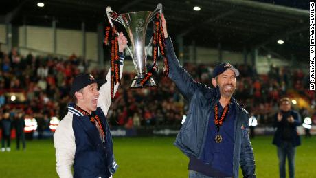 Rob McElhenney and Ryan Reynolds celebrate with the trophy on the pitch. 