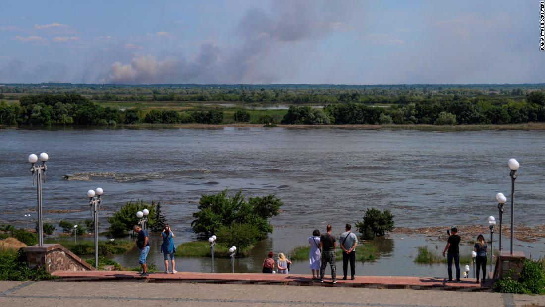 Local residents stand on an embankment of the flooded Dnipro River as smoke rises from shelling on the opposite bank.